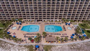 A hotel with two large swimming pools surrounded by sun loungers and umbrellas, a beach area with palm trees, and a high-rise building in the background. - AVAILABLE:4/12-4/19/895 /7 night/Couples Retreat/ balcony over looking beach (Panama City Beach)
