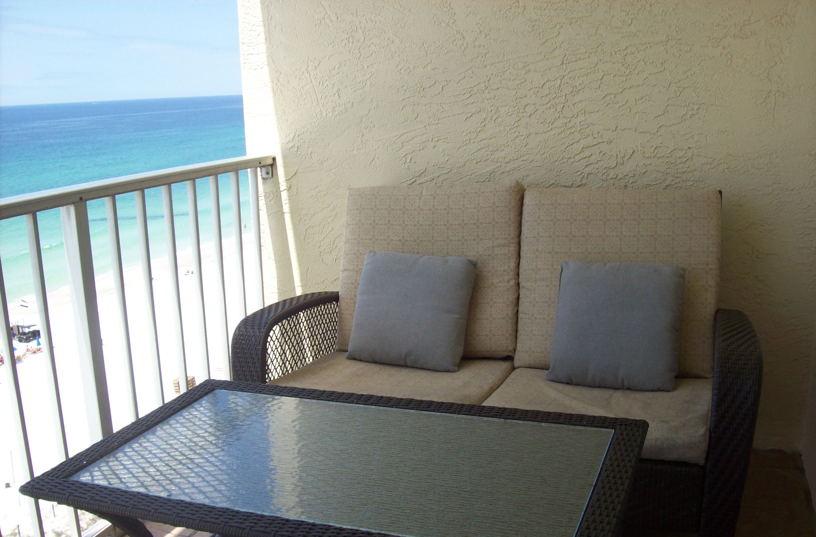 A balcony with a wicker sofa and a glass table, overlooking the ocean.