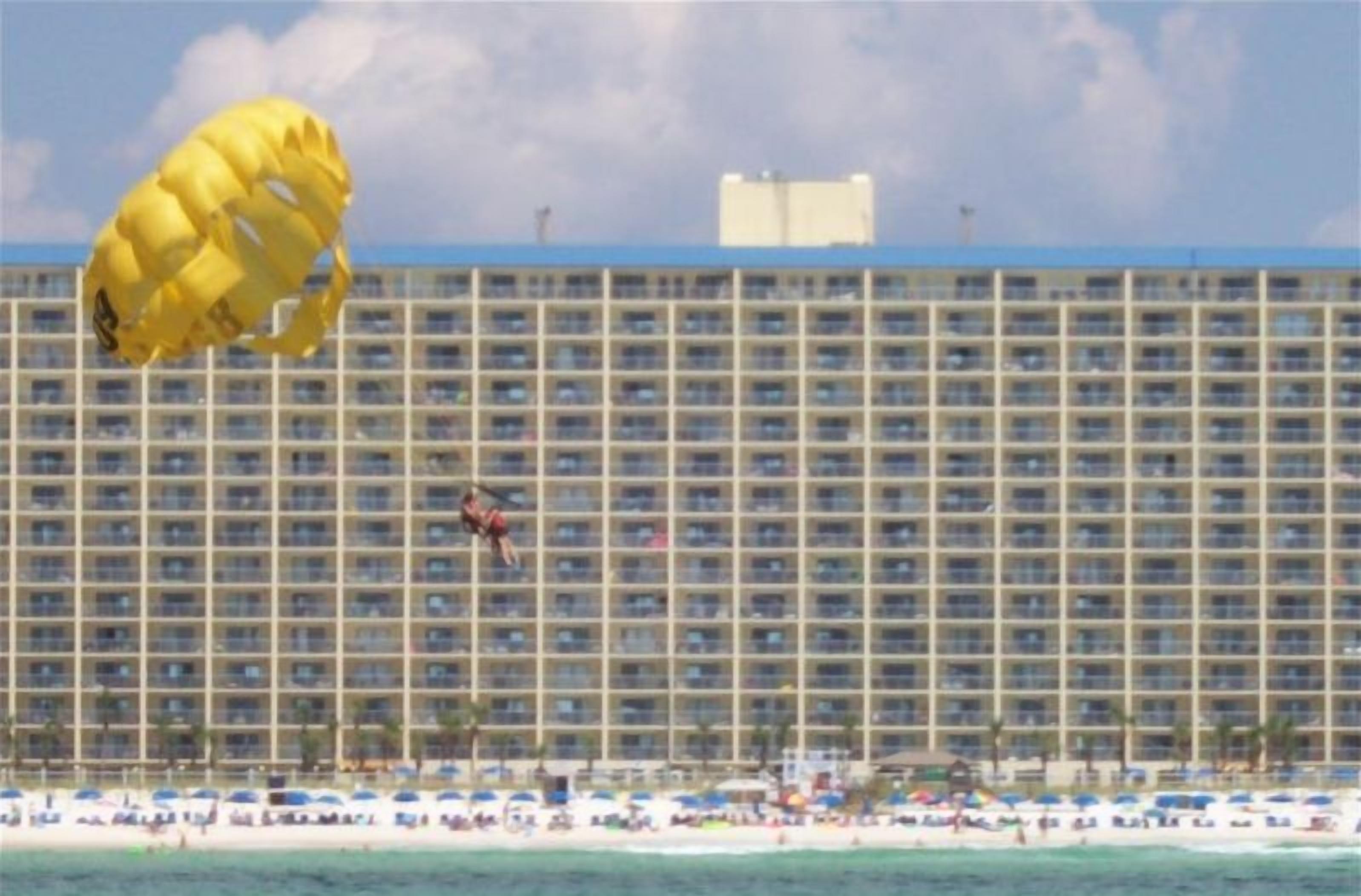 A hotel with a large building facade, a beach in the foreground, and a person parachuting.