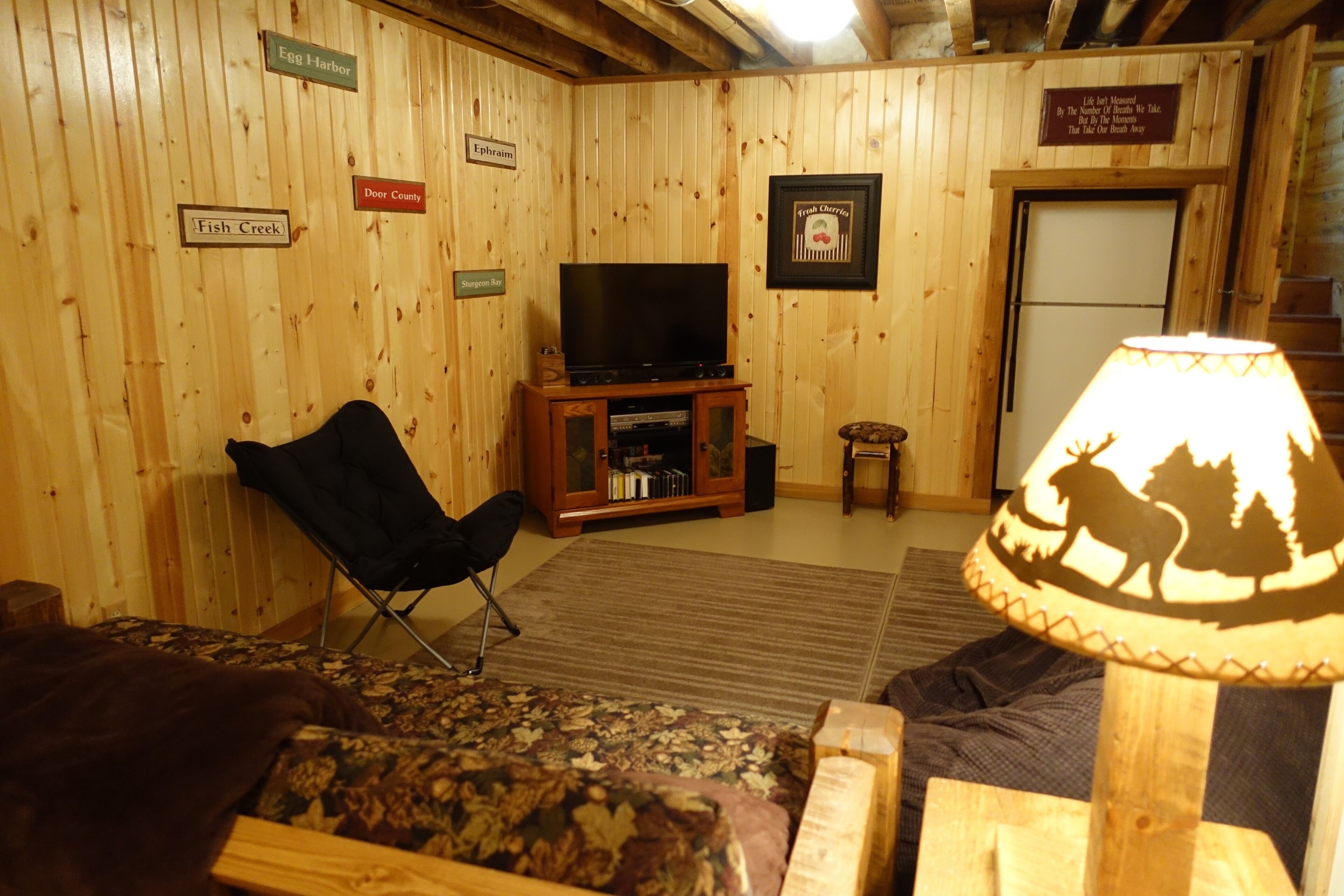 Ein gemütliches Zimmer mit Holzwänden, einem Flachbildfernseher auf einem Holzbodenständer, einem schwarzen Sessel, einem gemusterten Sofa und einer Lampe mit Waldmotiv.