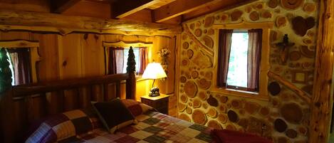 A cozy wooden cabin room with a bed, a nightstand, a lamp, and a stone wall with circular patterns.