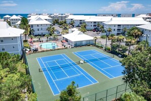 A tennis court with a pool, surrounded by white buildings with a view of the ocean.