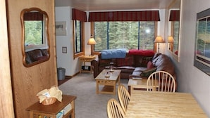 A cozy living room with a wooden dining table, a sofa, and two chairs. There are two lamps on the table and a window with a view of trees.
