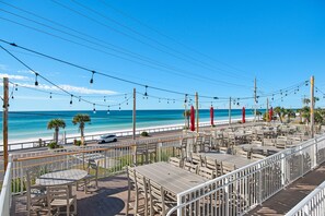 Un espace extérieur aménagé avec des tables et des chaises, offrant une vue sur une plage, un ciel bleu dégagé et l’océan.