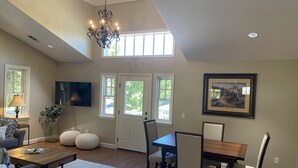 A spacious living room with a high ceiling, a chandelier, a dining area with a table and chairs, a television mounted on the wall, and a large window with a view of greenery outside.