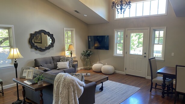 A spacious living room with a high ceiling, a chandelier, a flat-screen TV, a dining area with a table and chairs, and a large window providing natural light.