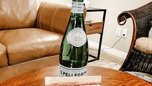 San Pellegrino -vesipullo, tervetulotoivotus ja paketti Altoids-minttupastilleja puisella pöydällä olohuoneessa, jossa on nahkasohva ja rottinkituoli.