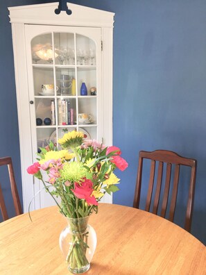 Une table à manger ornée d’un vase de fleurs, un buffet blanc aux portes en verre et deux chaises en bois.