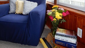 Un fauteuil bleu agrémenté de coussins à carreaux, une petite table en bois sur laquelle sont posés un vase de fleurs et des livres, et, par la fenêtre, une vue sur une clôture blanche.