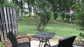 Un patio al aire libre con dos sillas de mimbre negro y una mesa de metal, rodeado por una valla de madera y un césped con árboles y un edificio al fondo.