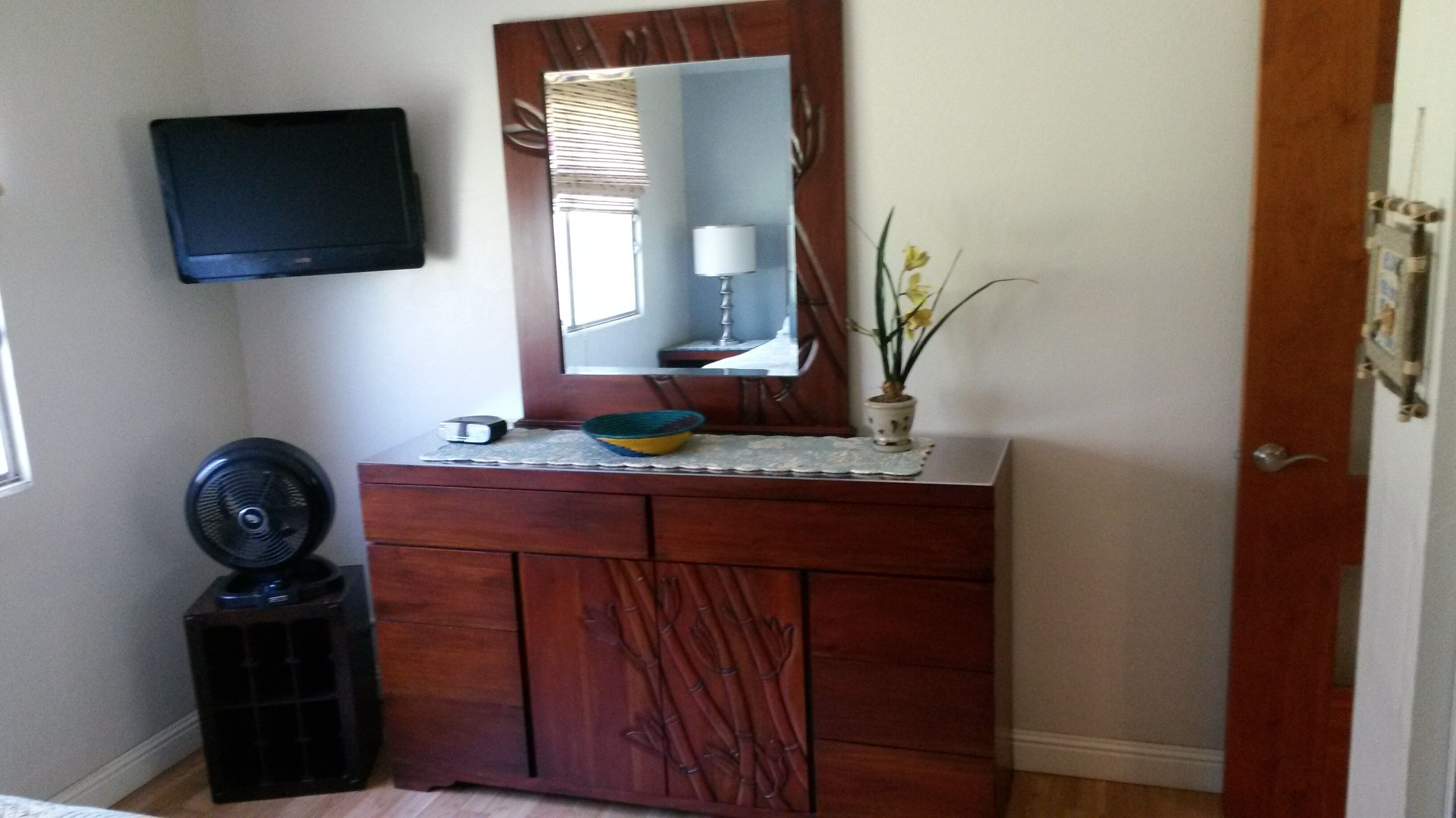 A room with a wooden dresser, a flat-screen TV mounted on the wall, a small fan, and a potted plant.