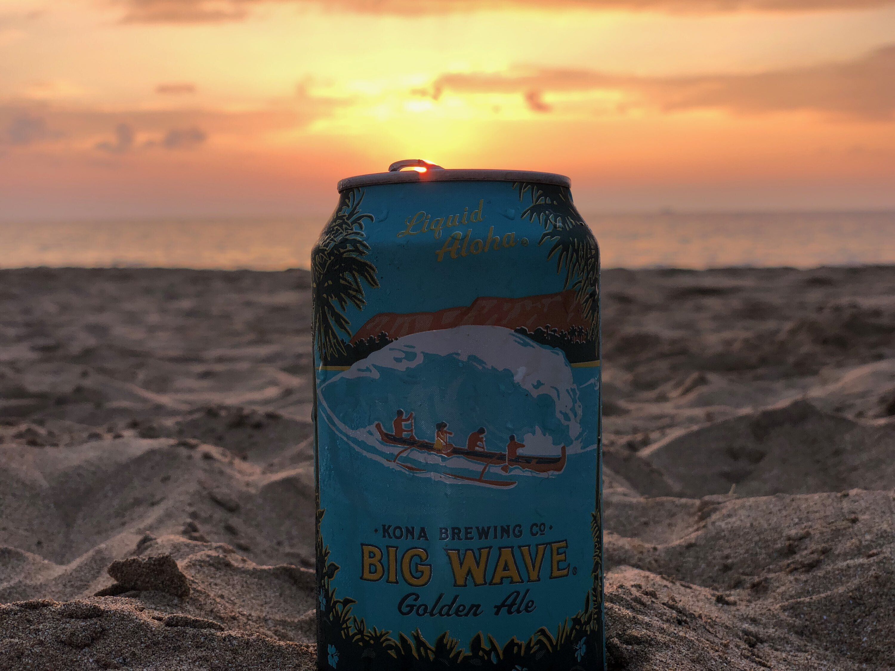 A can of Big Wave Golden Ale on a sandy beach with a sunset in the background.