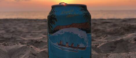 A can of Big Wave Golden Ale on a sandy beach with a sunset in the background.
