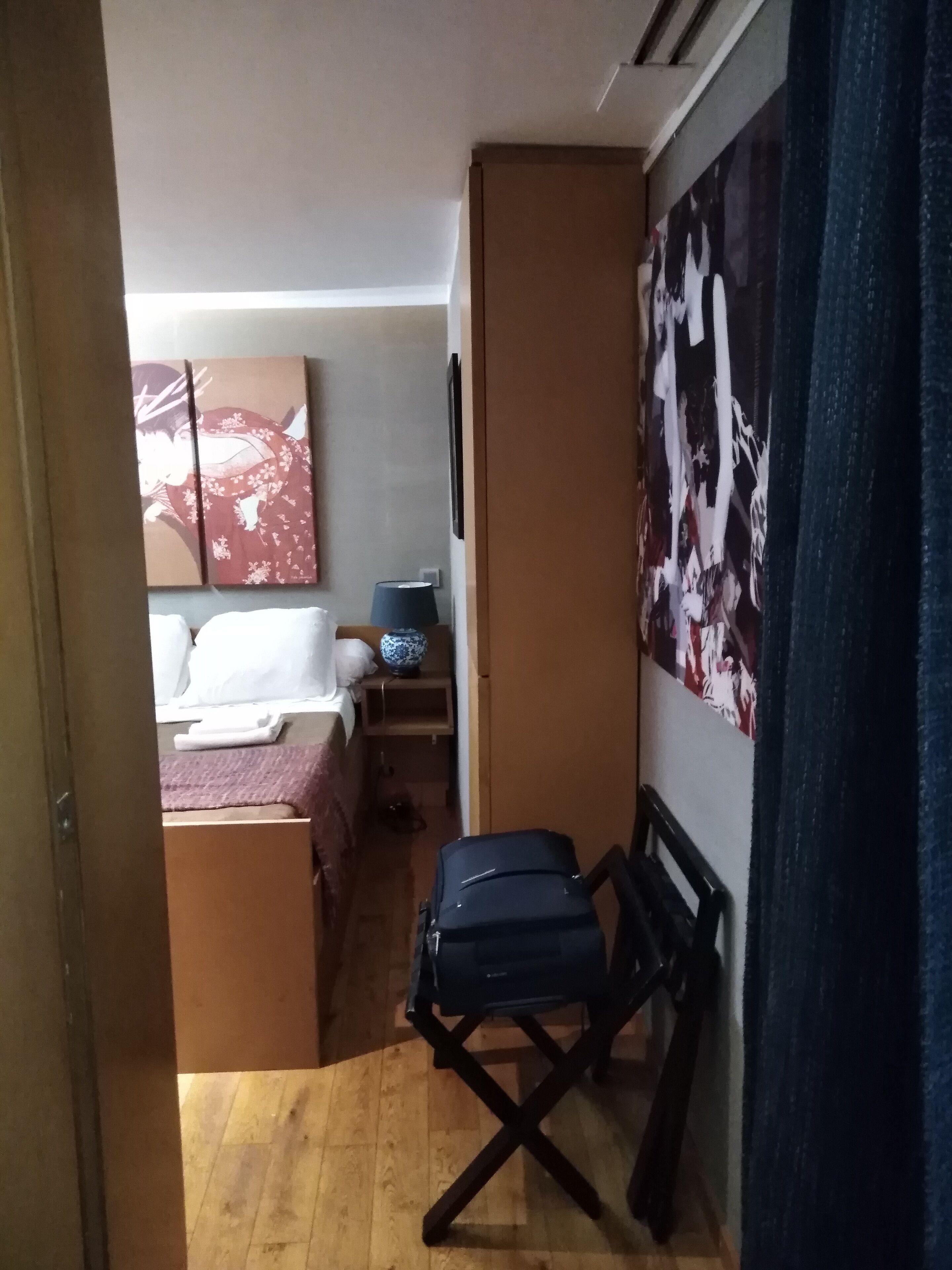 Een kleine kamer met een bed, een tafeltje met een lamp, een koffer en een muur met een groot abstract schilderij.