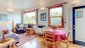 A cozy living room with a sofa, armchair, and a dining table with chairs. There are windows with curtains, a radiator, and a door with a handle.