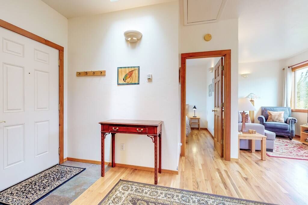 A hallway with a wooden floor, a red table, and a rug. There is a door on the left and a room visible in the background.