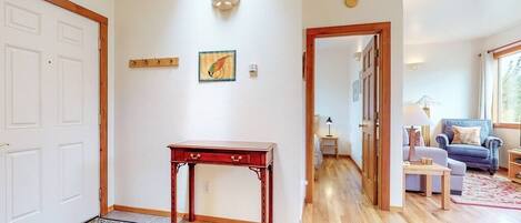 A hallway with a wooden floor, a red table, and a rug. There is a door on the left and a room visible in the background.