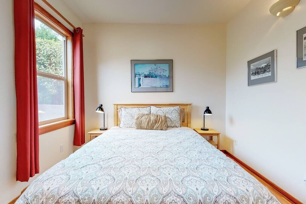 A bedroom with a large bed, two bedside tables, a window with red curtains, and framed pictures on the wall.