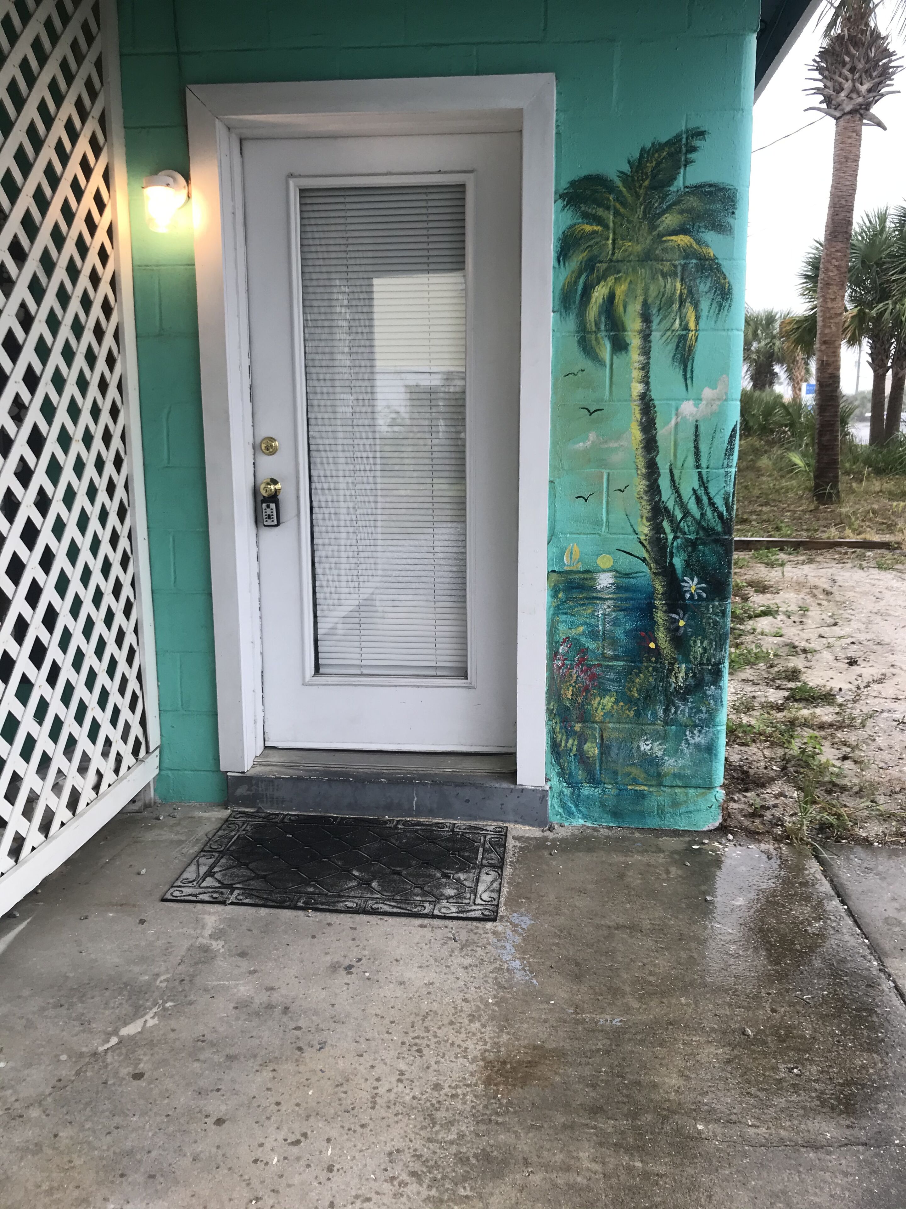 A door with a white frame and a window with horizontal blinds, a white lattice on the left, and a mural of a palm tree and flowers on the right.