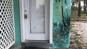 En dörr med en vit karm och ett fönster med horisontella persienner, ett vitt spaljé på vänster sida och en väggmålning med ett palmträd och blommor på höger sida.