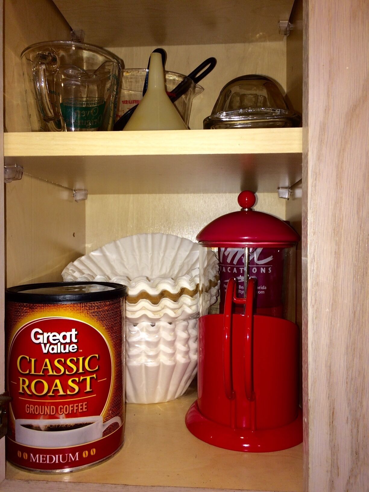 A kitchen shelf with a can of Great Value Classic Roast ground coffee, a white ceramic plate, and a red coffee pot.