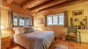 A cozy bedroom with a wooden ceiling, a bed with pillows, a nightstand, a chair, a bookshelf, and a window with curtains.