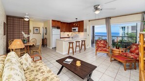 A spacious living room with a sofa, a coffee table, and a dining area with chairs and a table. There is a kitchen area with wooden cabinets and a refrigerator. The room has a balcony with a view of the ocean.