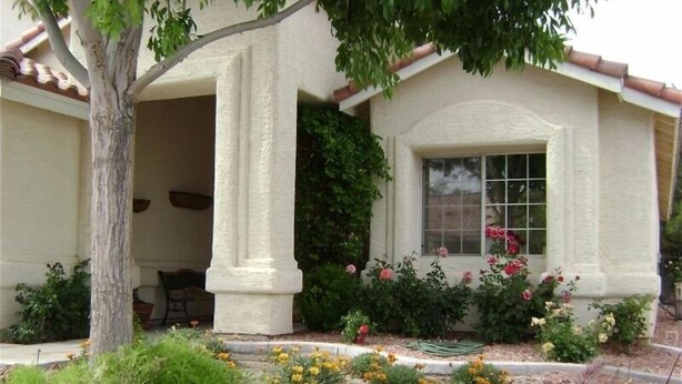 Una casa con porche cubierto, una ventana adornada con flores y un jardín con diversas plantas y flores.