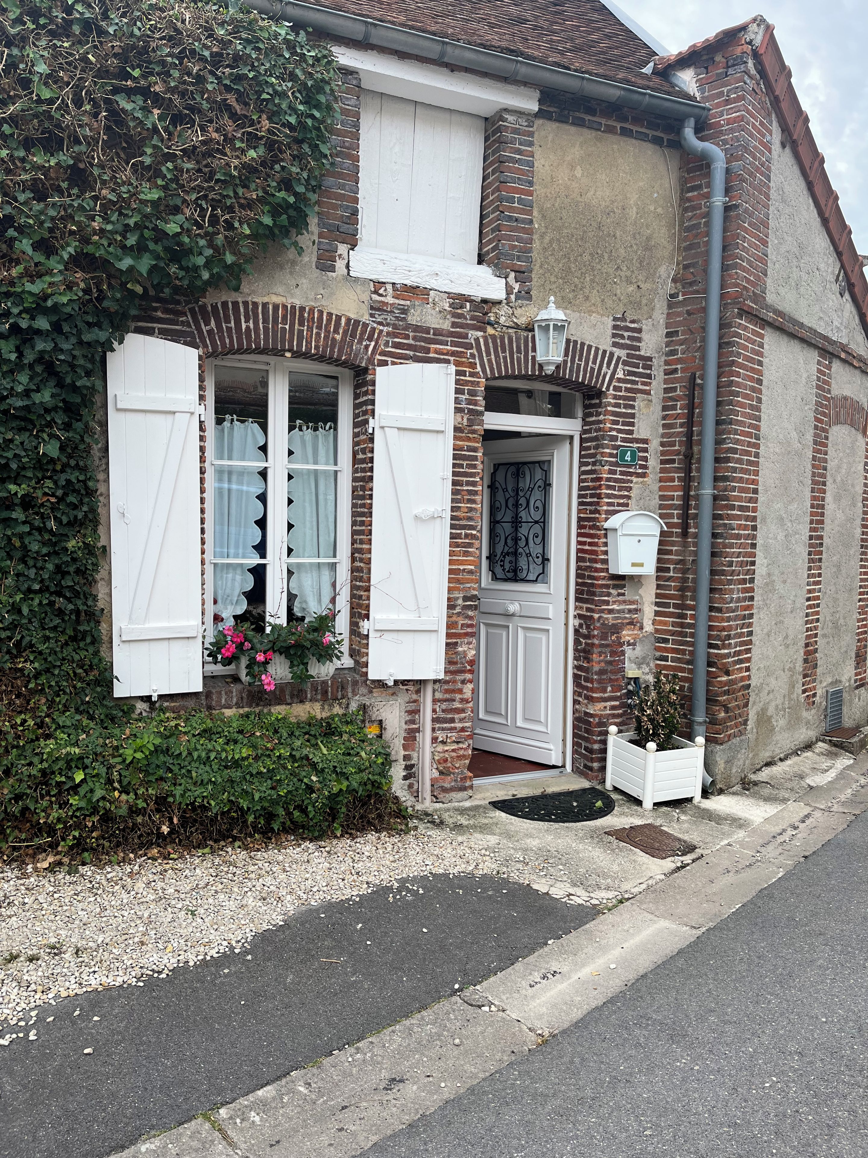 Ein zweistöckiges Haus mit Backsteinfassade, weißen Fensterläden und einem kleinen Garten mit rosa Blumen.