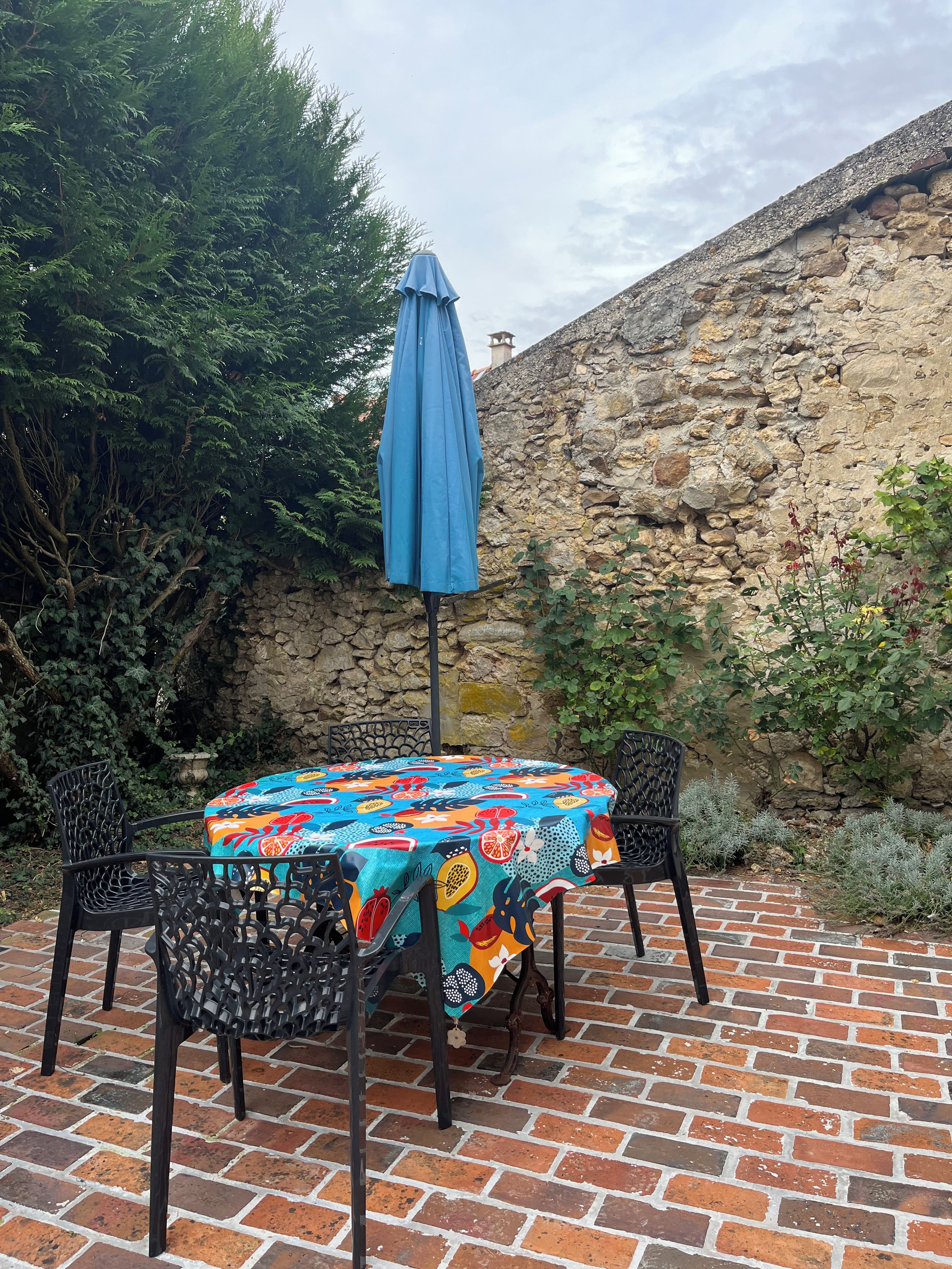 Eine Terrasse mit einem runden Tisch, der mit einer bunten Tischdecke bedeckt ist, zwei Stühlen und einem blauen Sonnenschirm.