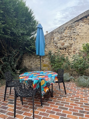 Eine Terrasse mit einem runden Tisch, der mit einer bunten Tischdecke bedeckt ist, zwei Stühlen und einem blauen Sonnenschirm.