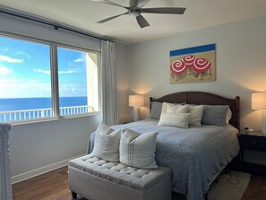 Une chambre à coucher comprenant un lit, une table de chevet, une lampe, un ventilateur de plafond, une fenêtre donnant sur l’océan et un tableau accroché au mur.