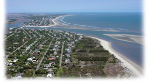 Vista aérea de uma área residencial costeira, com casas ao longo da praia, céu claro e uma estrada visível que leva à praia.