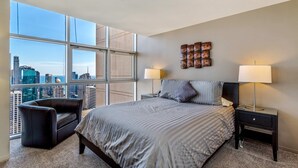 A bedroom with a large bed, two bedside tables with lamps, a chair, and a city view through large windows. - 55th-Floor 3BR Duplex Penthouse  Lake & City Views (Chicago)