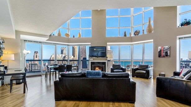 A spacious living room with a large black leather sofa, a television, and a fireplace. The room features large windows with a city skyline view. - 55th-Floor 3BR Duplex Penthouse  Lake & City Views (Chicago)