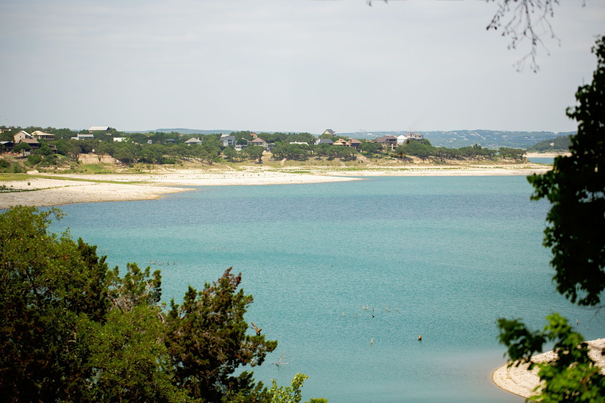 En kustvy med klart blått vatten, en sandstrand och hus i bakgrunden.