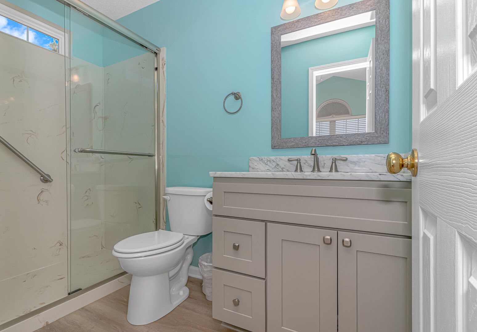 A bathroom with a glass shower enclosure, a toilet, a sink with a marble countertop, and a mirror with a decorative frame.