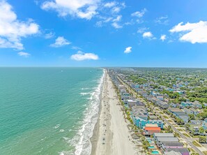 Vue aérienne d’une ville côtière avec une plage, des immeubles résidentiels et un ciel bleu dégagé.