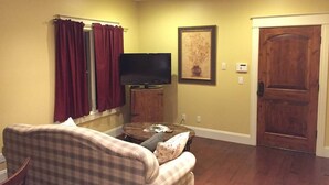 A living room with a plaid sofa, a flat-screen TV, a wooden door, and a painting on the wall. - Tuscan Villa with Private Courtyard in Briarbrook (near Joplin Regional Airport) (Carl Junction)