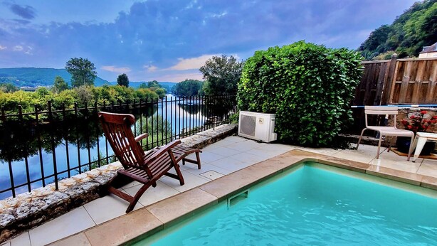 A pool with a wooden chair and a table, surrounded by a stone wall and a metal fence, overlooking a river and greenery.