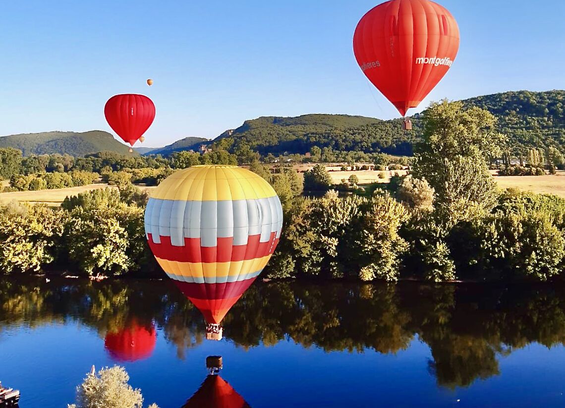 A hot air balloon over a body of water with reflections, surrounded by trees and hills.