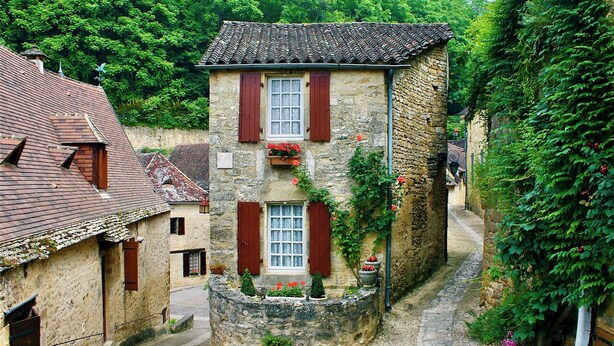 Une ruelle étroite pavée, bordée de bâtiments en pierre aux volets rouges, un mur de pierre avec un petit jardin, et une végétation luxuriante en arrière-plan.