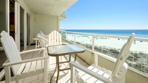 A balcony with white Adirondack chairs and a small table, overlooking a beach with umbrellas and a clear sky.