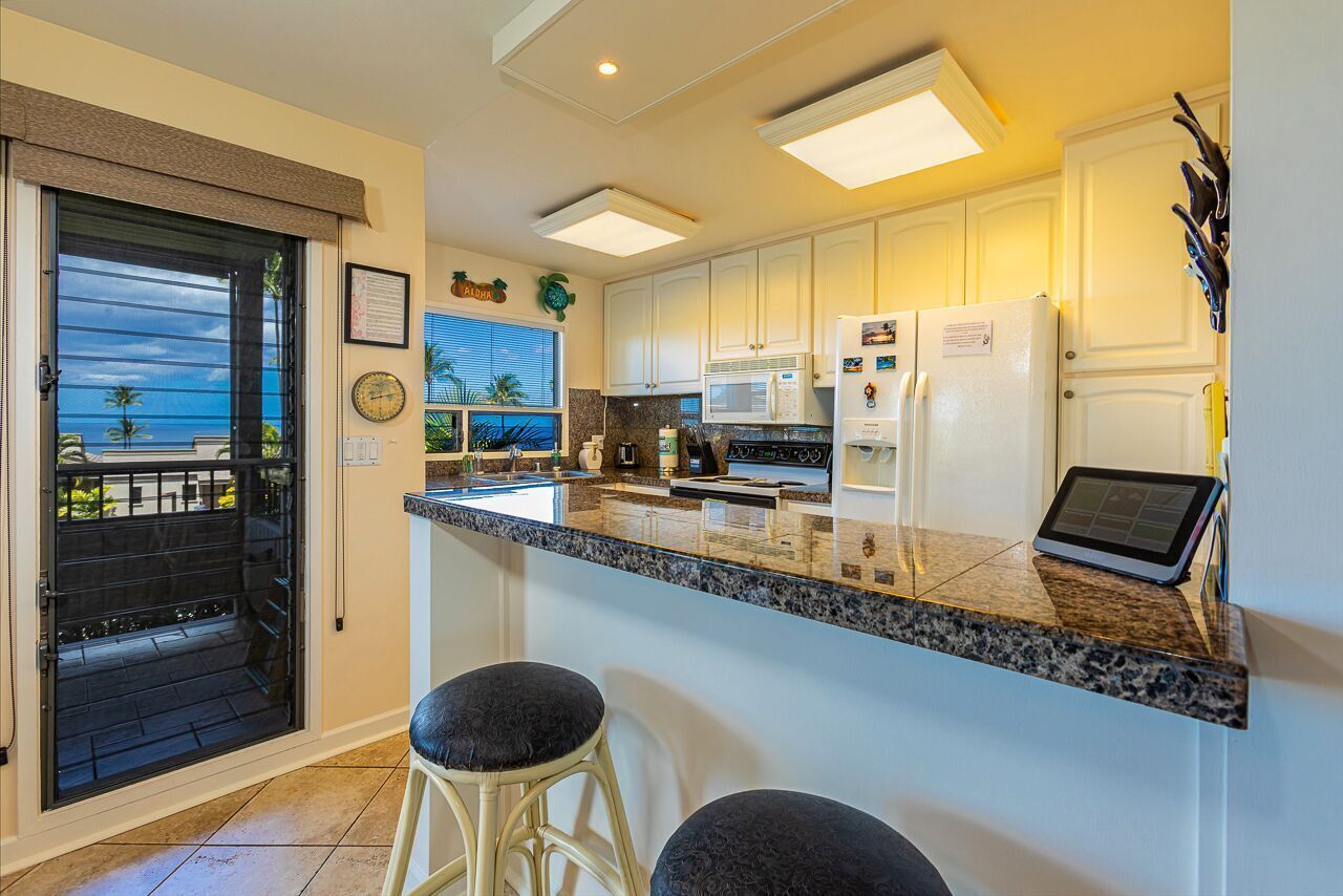 Una cocina moderna con armarios blancos, encimera de granito y refrigerador empotrado. Hay dos taburetes de bar y una ventana con vistas a palmeras.