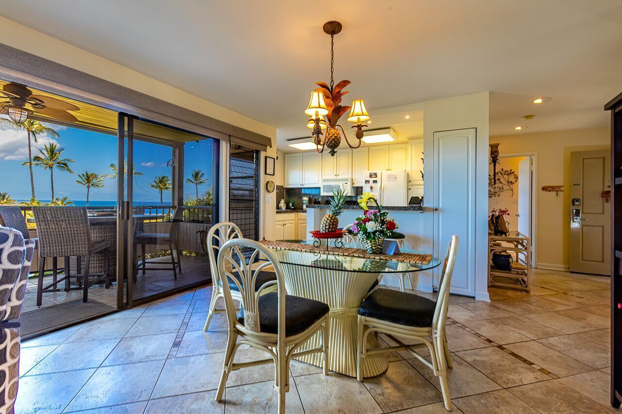 A dining area with a glass table and wicker chairs, a kitchen with white cabinets, and a view of palm trees and the ocean.