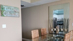 Un comedor con una mesa de vidrio y sillas tejidas, un letrero en la pared y un espejo grande que refleja el espacio.