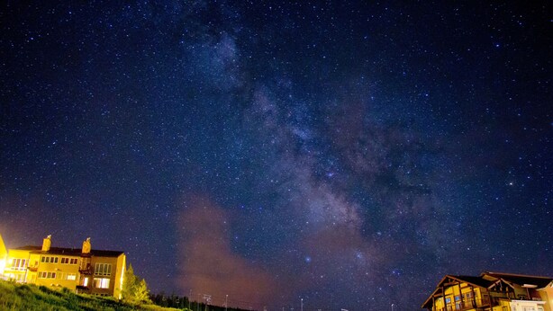 A night sky with stars and the Milky Way over a grassy hill with two buildings.