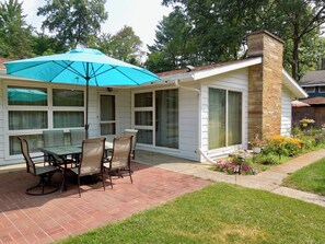 Eine überdachte Terrasse mit Tisch und Stühlen, ein Backsteinboden und ein Garten mit Blumen.