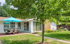 A covered patio with a table and chairs, a blue umbrella, and a brick pathway leading to a house with a brick exterior. - All Season Lakehouse on Private Lake in Southwest Michigan (Gobles)