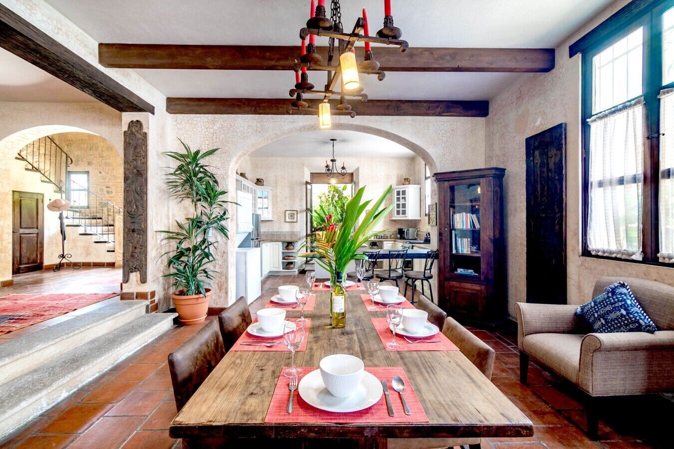 Un comedor con una mesa de madera dispuesta para seis personas, rodeada de sillas. Se ve una escalera que sube a un nivel superior, una cocina al fondo y una estantería a la derecha.